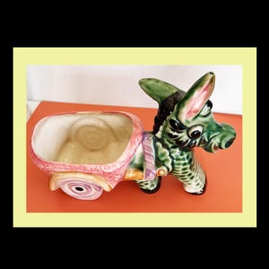 May include: A ceramic donkey planter with a cart. The donkey is green with black and pink accents. The cart is pink and beige with a purple strap. The planter is a decorative item.