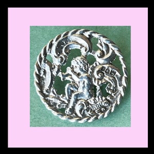 May include: A silver-toned metal button with a detailed design of a cherub playing a musical instrument within a circular frame.