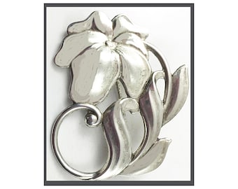 May include: A polished silver-toned metal brooch shaped like a single flower with five rounded petals. The design includes a curved stem that forms a circular loop at the base and two elongated, smooth leaves extending to the right side.