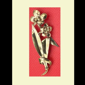 May include: A gold-toned metal brooch in the shape of a stylized flower arrangement. The brooch features two flowers, leaves, and a curved stem. The background is a textured red surface.