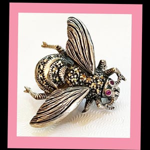 May include: A detailed silver-toned bee brooch with marcasite detailing and ruby-colored accents. The wings are textured with etched lines. The brooch is set against a white background with a pink border.