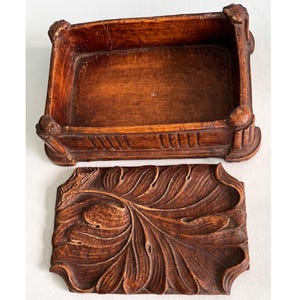 May include: A rectangular wooden box with a lid. The box and lid are a warm brown color. The box has decorative corner posts and carved details. The lid is carved with a large leaf design. The box is open.