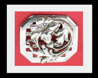May include: A silver-toned brooch with a floral design. The brooch features a central flower surrounded by leaves and is set within an octagonal frame. The background is a textured red.