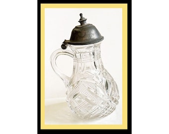 May include: A clear glass pitcher featuring a faceted geometric pattern and a curved handle. The vessel is topped with a dark, weathered metal lid that includes a small, pointed finial and a hinged mechanism attached to the handle.