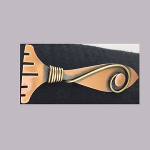 May include: A copper and brass brooch in the shape of a stylized fish. The fish has a curved tail and a body with a textured surface. The brooch is attached to the fish's body with a wire loop.