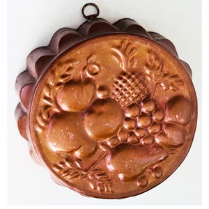 May include: A copper-colored, round mold with a decorative fruit design. The mold features a pineapple, grapes, apples, and pears. It has a scalloped edge and a small ring for hanging. This kitchenware item is likely used for baking.