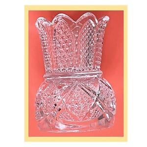 May include: A clear glass vase featuring a scalloped rim and a complex geometric pattern. The body displays a combination of diamond-shaped facets and cross-hatched textures, creating a reflective surface that catches light across its entire structure.