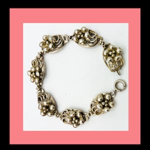 May include: Silver-toned bracelet featuring a floral design. The bracelet has multiple oval-shaped links, each adorned with clusters of small, round beads and intricate floral details. A circular clasp secures the bracelet.