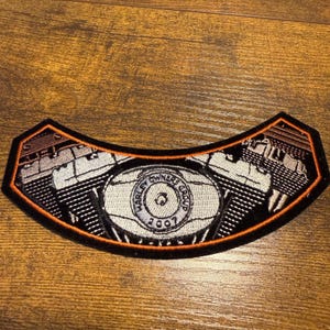 May include: A curved embroidered patch featuring a detailed silver and black motorcycle engine graphic. The center circular emblem displays the text "HARLEY OWNERS GROUP" and "2007" in black lettering. The patch has a thick black border with an orange accent line.