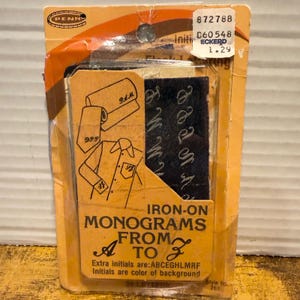 May include: A package of iron-on monograms from A to Z featuring dark blue fabric letters with white script embroidery. The orange cardboard packaging displays illustrations of a tie and shirt with monograms, along with the text Iron-On Monograms From A To Z.