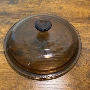 May include: A round, transparent amber-colored glass lid for cookware, featuring a centered black circular knob handle. The rim of the lid is embossed with the text PYREX V-15-C, indicating the brand and model identification for this kitchen item.
