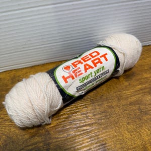 May include: A skein of off-white Red Heart sport yarn with a paper label featuring the brand logo, a heart icon, and the text Machine Washable & Dryable. The yarn is wound into a cylindrical shape with a soft, textured appearance and matte finish.