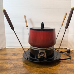 May include: A vintage red and black Cornwall fondue pot with four fondue forks. The pot has a black base and lid, with a red body. The forks have wooden handles with colored tips. The control dial reads "Cornwall".