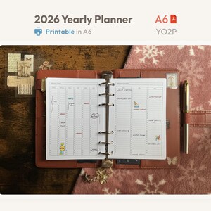 May include: An open A6 2026 yearly planner with a brown leather-like cover and a metal ring binder mechanism. The white pages feature a vertical schedule layout with handwritten notes and small colorful stickers. A gold pen is tucked into the side.