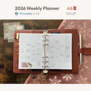 Puede incluir: Una agenda semanal A6 de cuero marrón con anillas, abierta en una página de calendario con cuadrícula, notas manuscritas y pegatinas. Incluye un mecanismo metálico de seis anillas, un bolígrafo dorado y el texto 2026 Weekly Planner, A6 y WO2P.