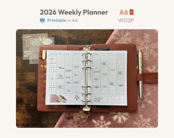Könnte beinhalten: Ein brauner A6-Ringplaner aus Leder, aufgeschlagen auf einer Kalenderseite mit Raster, handschriftlichen Notizen und Aufklebern. Der Planer verfügt über eine Metall-Ringmechanik, einen goldenen Stift und den Text 2026 Weekly Planner, A6 und WO2P.
