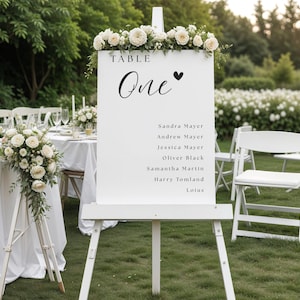 Puede incluir: Un letrero rectangular blanco de distribución de mesas de boda exhibido en un caballete de madera blanca. El letrero presenta el texto TABLE One con un icono de corazón y una lista de nombres que incluye a Sandra Mayer, Andrew Mayer, Jessica Mayer, Oliver Black, Samantha Martin, Harry Tomland y Loius, coronado con arreglos florales blancos.