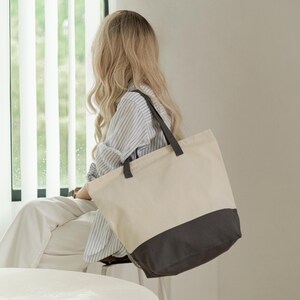 A woman with long blonde hair is sitting in a room, looking out a window. She is wearing a striped shirt and carrying a large tote bag.