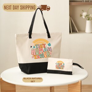 May include: A canvas tote bag and matching pouch set. The tote bag is off-white with a black bottom and handles. The pouch is also off-white with a black bottom. Both feature a colorful graphic that reads "TINY HUMAN TAMER".