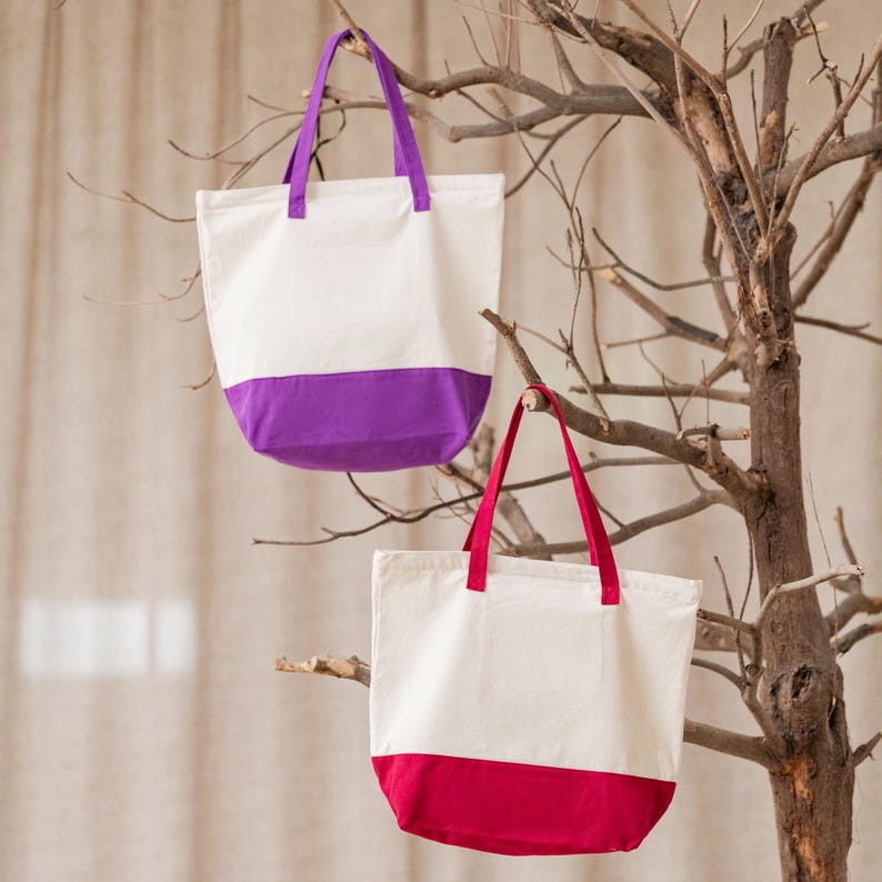 Two colorful tote bags, one white and purple, the other red and white, are hanging from a tree branch.