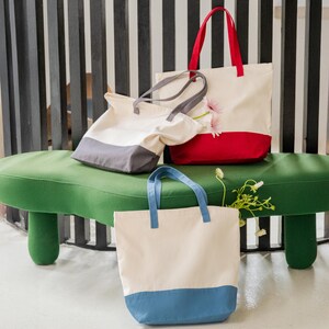Three colorful tote bags are displayed on a green bench, with one red and white bag and two gray and white bags.