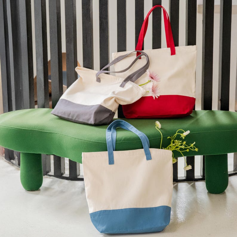 Three colorful tote bags are displayed on a green bench, with one red and white bag and two gray and white bags.