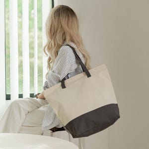 A woman with long blonde hair is sitting in a room, looking out a window. She is wearing a striped shirt and carrying a large tote bag.