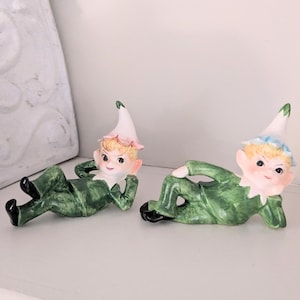 May include: Two ceramic elf figurines in a relaxed pose. They wear green outfits, black shoes, and white hats with pink and blue accents. The elves have blonde hair and rosy cheeks, adding a whimsical touch to the decor.