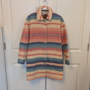 Puede incluir: Un abrigo largo con botones con un diseño de inspiración suroeste. El abrigo presenta rayas horizontales en tonos melocotón, azul y crema, con un canesú estampado. Tiene cuello y dos bolsillos delanteros. El abrigo está colgado de una percha blanca.