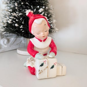 May include: A vintage Christmas figurine of a child in a red and white outfit, wrapping a gift. The figurine has a serene expression and is kneeling beside a stack of white gift boxes decorated with green trees and the word "R MAS".