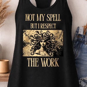 Puede incluir: Camiseta sin mangas negra con el texto "NOT MY SPELL BUT I RESPECT THE WORK" y un gráfico de una mujer con cabello de serpiente. La camiseta sin mangas está colgada en una percha de madera.