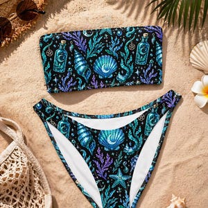 May include: A black two-piece bikini set featuring a repeating pattern of teal and purple seashells, potion bottles, and seaweed. The bandeau-style top and high-waisted bottoms create a striking beachwear ensemble.