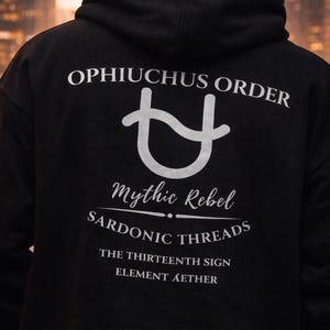 May include: Black hoodie featuring white text: "Ophiuchus Order," a symbol, "Mythic Rebel," "Sardonic Threads," "The Thirteenth Sign," and "Element Aether." The hoodie has a hood and is a dark colour, with a blurred background.