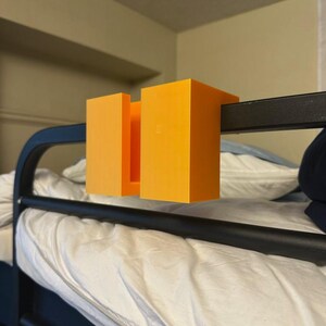 May include: Bright orange, 3D-printed bunk bed accessory. The item is designed to attach to the black metal frame of a bunk bed. The accessory is composed of three rectangular blocks, two outer and one inner.