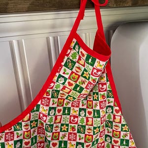 May include: A festive apron featuring a repeating chequered pattern of red, white, and green squares. Each square contains small holiday-themed illustrations including snowmen, Christmas trees, stars, stockings, bells, and rocking horses.