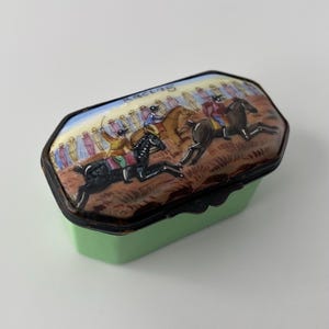 May include: A small, rectangular, light green trinket box with a hinged lid. The lid features a hand-painted scene of a horse race with jockeys, horses, and spectators, including the word "Races" written in script at the top of the lid.