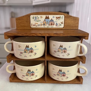 May include: A set of four cream-coloured soup bowls with handles, each decorated with a quaint village scene. The bowls are displayed in a wooden rack with a decorative tile featuring a similar pastoral design. The rack is made of light brown wood.