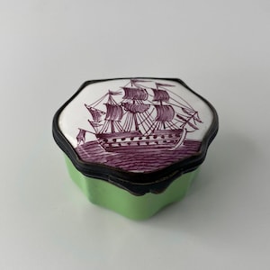 May include: A hexagonal trinket box with a light green base and a dark metal rim. The lid features a white surface with a detailed purple illustration of a sailing ship on the water. The box has a scalloped edge design around the perimeter.