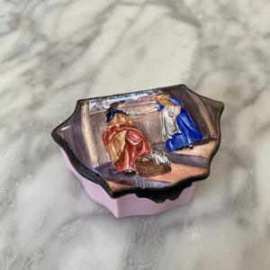 May include: A small, scalloped-edge trinket box with a light pink base and a hinged lid. The lid features a hand-painted enamel scene of two figures in period clothing, one in a red dress and one in a blue dress, with the text New Mackerel New.