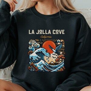 May include: Black sweatshirt featuring a graphic of a sunset over ocean waves, a flying bird, and the text "LA JOLLA COVE California". The design incorporates shades of blue, orange, and beige.