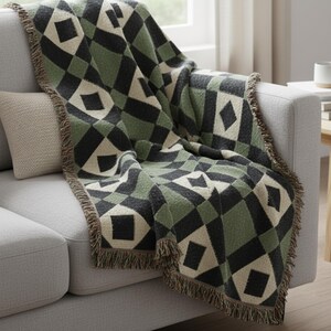 May include: A patterned throw blanket in green, black, and cream, draped over a grey sofa. The blanket has a diamond design and fringed edges, adding texture and style to the living space. The sofa is next to a window.