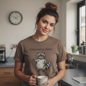May include: A brown t-shirt with the text "clinically feral" above a black and white raccoon illustration. The raccoon is holding a small object. The shirt has rolled-up sleeves.