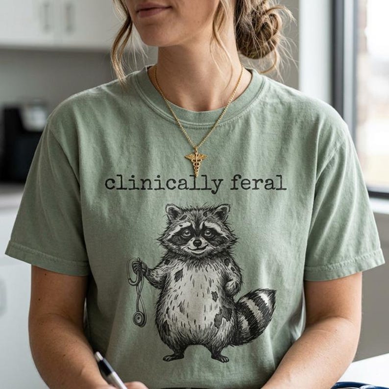 May include: Sage green t-shirt with the text "clinically feral" above a raccoon illustration holding a stethoscope. The raccoon is black and white with a striped tail. A gold necklace with a medical symbol is worn.