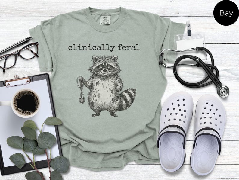 May include: Sage green t-shirt with the text "clinically feral" above a raccoon illustration holding a stethoscope. The image includes a stethoscope, white clogs, glasses, and a clipboard with a cup of coffee.