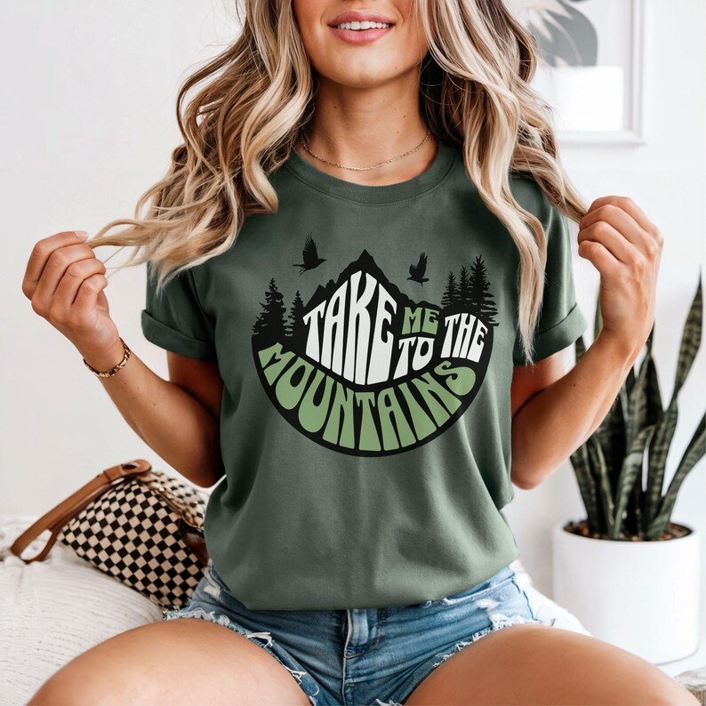 Puede incluir: Camiseta verde oliva con el texto "TAKE ME TO THE MOUNTAINS" en un dise&ntilde;o blanco y verde. El dise&ntilde;o incluye una silueta de monta&ntilde;a, &aacute;rboles y p&aacute;jaros. La camiseta tiene mangas cortas.