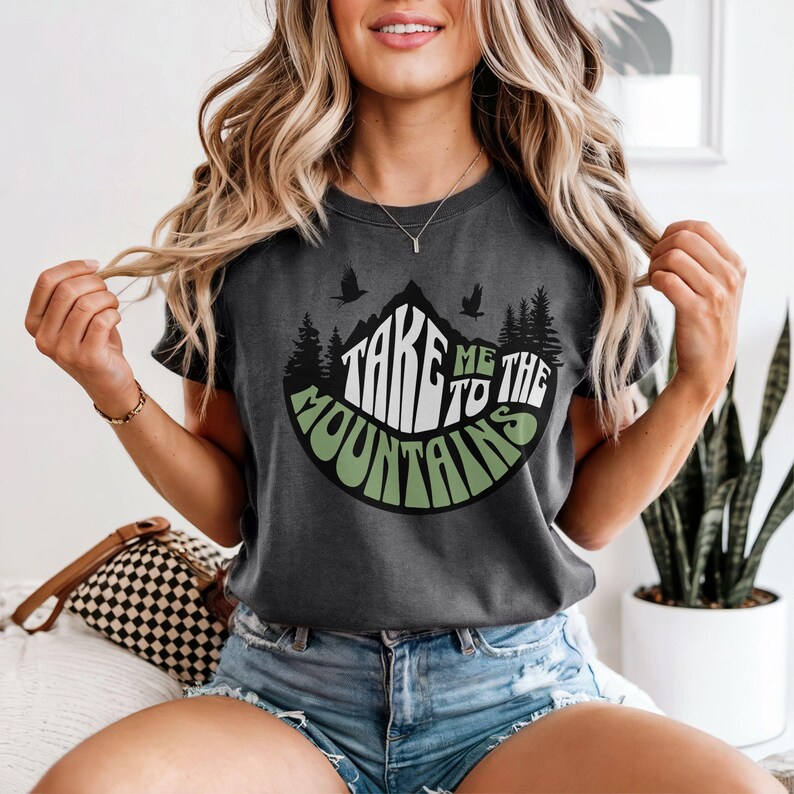 Puede incluir: Camiseta gris oscuro con el texto "TAKE ME TO THE MOUNTAINS" en blanco y verde. El dise&ntilde;o presenta un gr&aacute;fico de monta&ntilde;a con &aacute;rboles y p&aacute;jaros. La camiseta es de manga corta.