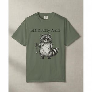 May include: Olive green t-shirt featuring a black and white illustration of a raccoon holding a pocket watch. The text "clinically feral" is above the raccoon graphic. The shirt has a crew neck and short sleeves.