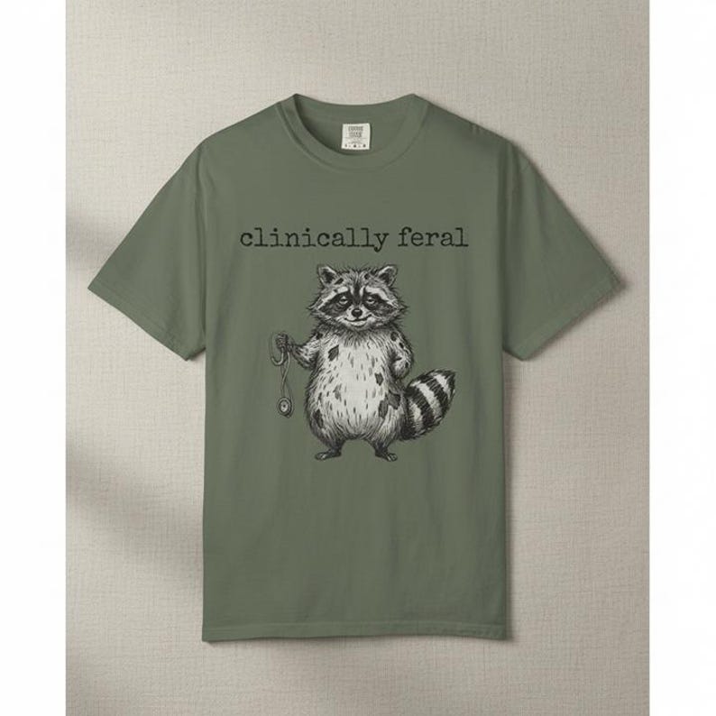 May include: Olive green t-shirt featuring a black and white illustration of a raccoon holding a pocket watch. The text "clinically feral" is above the raccoon graphic. The shirt has a crew neck and short sleeves.