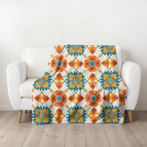 Op de afbeelding: Een witte bank met een sprei met een herhalend bloemenpatroon in oranje, teal en geel. De deken is over de bank gedrapeerd en toont het ingewikkelde ontwerp. De sprei voegt een vleugje kleur en patroon toe aan de kamer.
