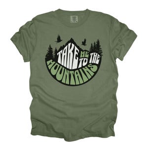 Puede incluir: Camiseta verde oliva con un dise&ntilde;o de monta&ntilde;a. El dise&ntilde;o presenta el texto "TAKE ME TO THE MOUNTAINS" en blanco y verde, con siluetas de &aacute;rboles y p&aacute;jaros. Una camiseta c&oacute;moda e informal.